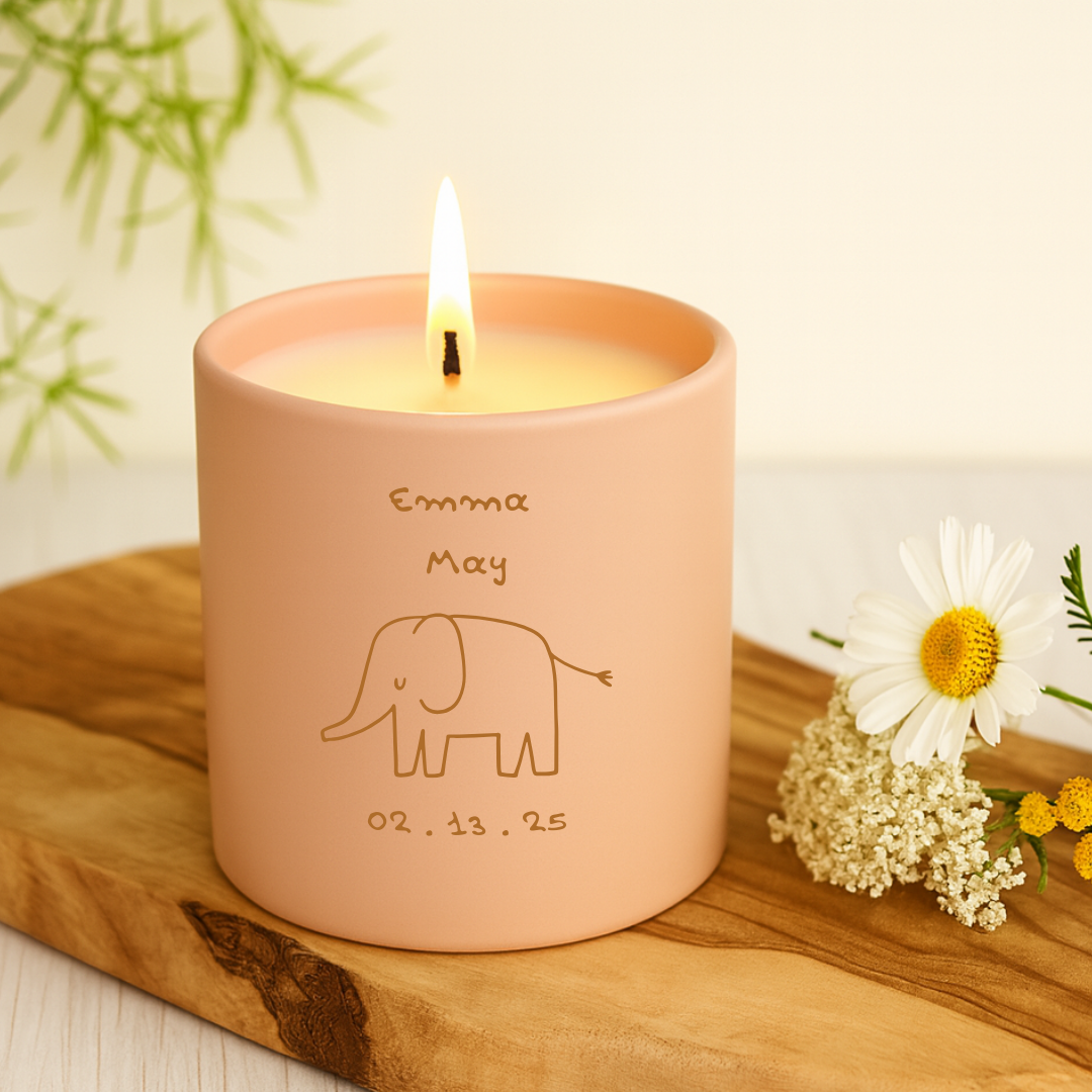 Pink candle with 'Emma May' and an elephant illustration on a wooden surface with flowers.