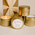 Gold candle tins with 'Wedding Cake' label and one lit candle on a beige background
