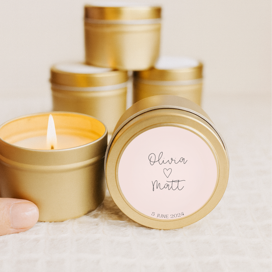 Gold candle tin with a lit candle, pink label with 'Olivia ❤️ Matt' text, on a light background.