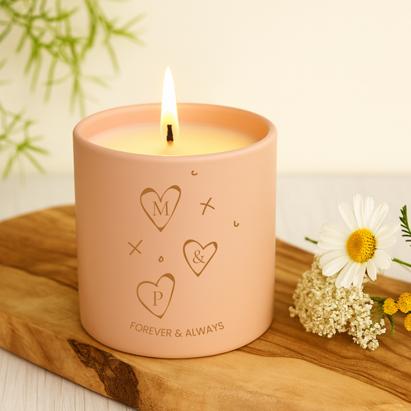 Pink candle with engraved heart symbols and initials on a wooden surface with flowers.