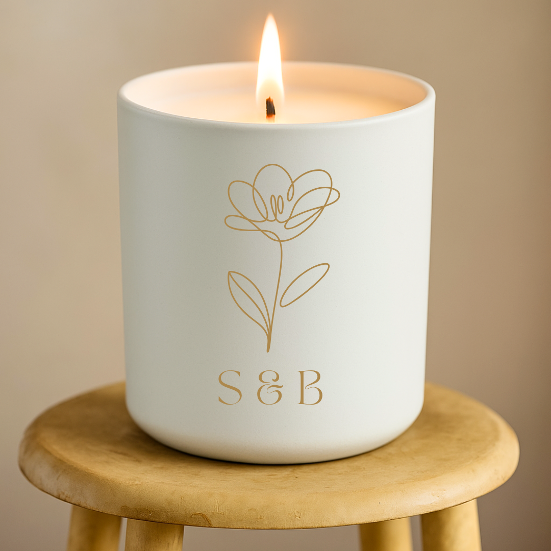 Candle in a white container with a floral design and 'S&B' initials on a wooden stool.