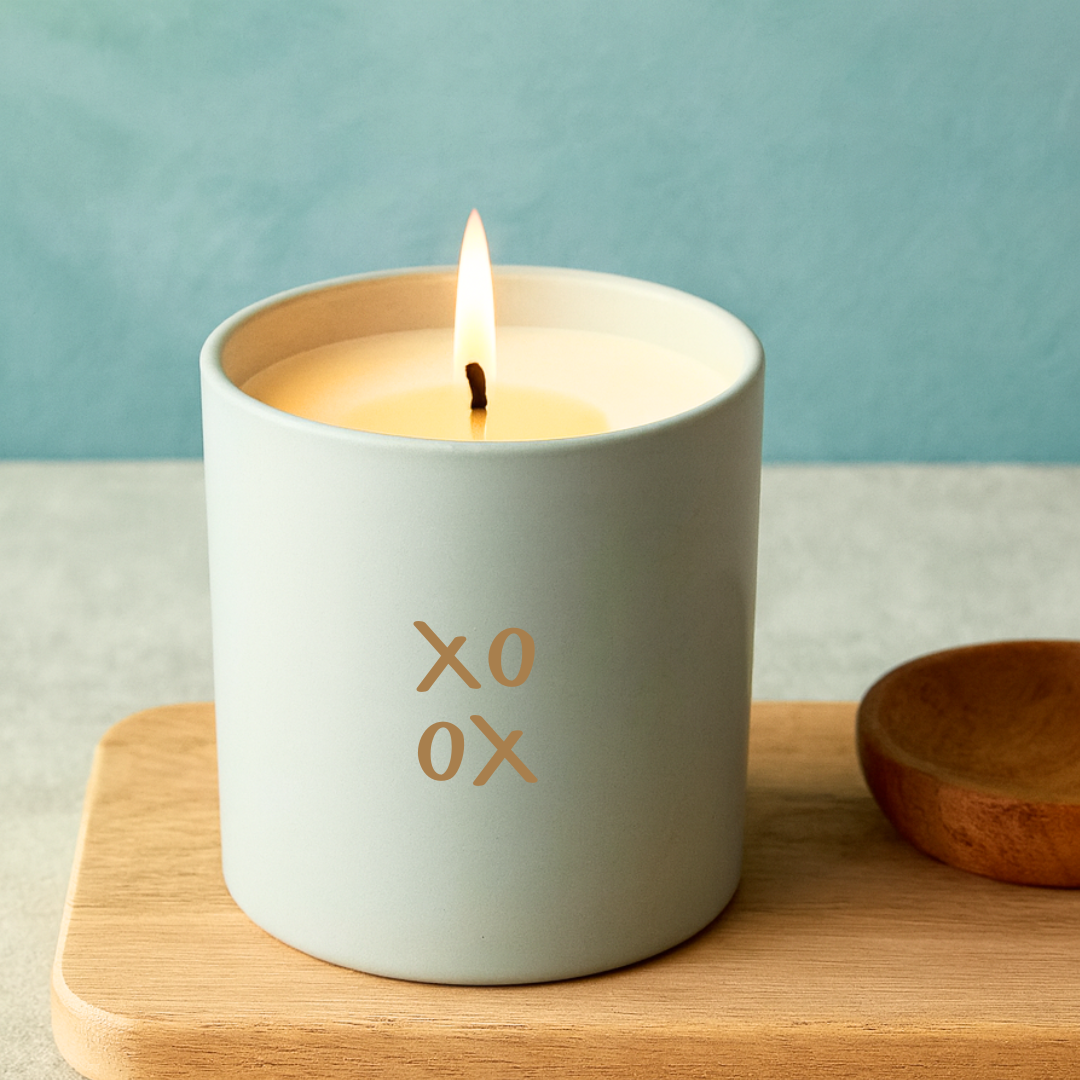 Candle in a white container with 'XO' on a wooden surface against a teal background