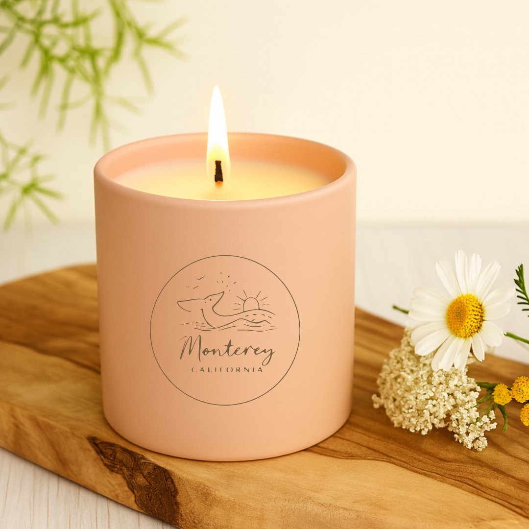 Pink candle with 'Monterey, California' branding on a wooden surface with flowers.